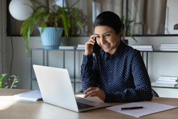 Smiling young indian woman sit at desk in office look at laptop screen talking on cellphone. Happy...