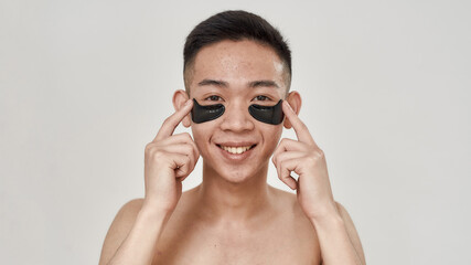 Portrait of shirtless young asian man applying black patches under the eyes, smiling at camera isolated over white background. Beauty, skincare routine, cosmetology for men