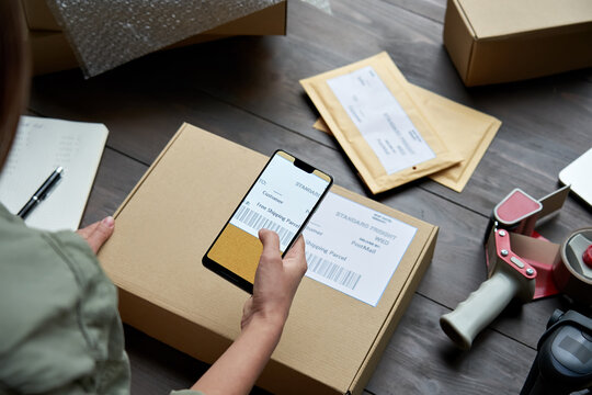 Female Warehouse Worker, Seller, Dropshipping Small Business Owner Holding Phone Scanning Retail Package Postal Parcel Barcode On Ecommerce Shipping Box Label On Smartphone Using Mobile App, Close Up.