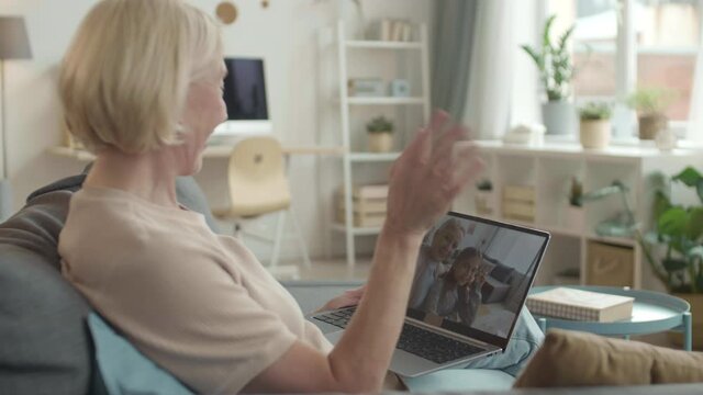 Over-the-shoulder shot of white-haired woman sitting in living room holding laptop on knee for video chatting with short-haired female relative and school girl