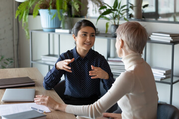 Motivated young multiracial female colleagues sit at desk in office talk shared thoughts discuss...