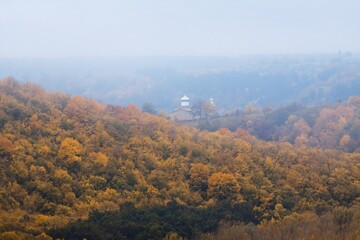 mountains covered with magnificent mountain forest, green, yellow and orange autumn leaves, cloudy and misty October morning, Orthodox church in poor visibility, ecotourism active rest concept