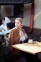 young blonde woman in fashionable dress, yellow plaid woolen jacket sits on the terrace of a cafe with coffee.