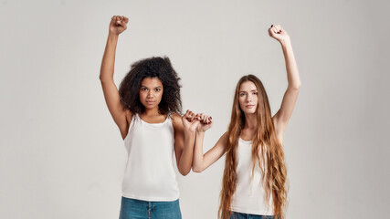 Portrait of two young diverse women, best friends looking serious at camera while raising arm and...