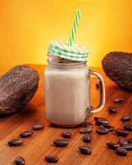 glass of cocoa drink with a green straw, acompanied by dried cacao fruits an cacao beans before an orange background. 