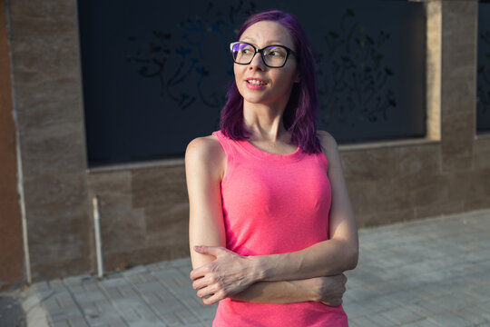 Young Caucasian woman has her attention drawn to one side, looks to be interested in what she sees. Dressed in pink casual clothes, with glasses and purple hair, basking in the golden hour light