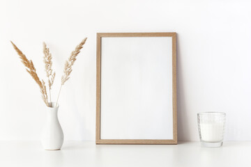Empty beige wooden frame stands on white table with dry grass in vase and candle. Mockup poster...