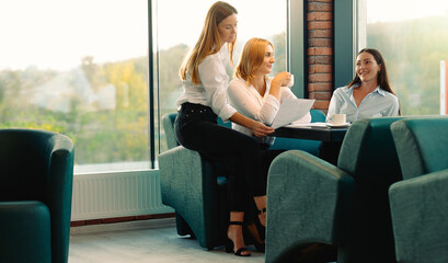 Fototapeta premium Friends having fun drinking coffee in modern office brainstorming under new project. Young women discuss sitting together at table spending time in coffee break.