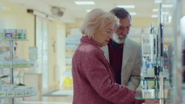 Close up elderly couple walking on drugstore. Choose pills. Feel headache. Conversation pharmaceutical client. Seller commercial health care buyer uniform. Slow motion - Powered by Adobe