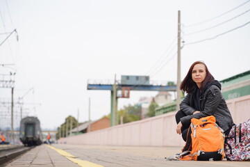 Young woman squats on platform, waiting for train. Female passenger with backpacks sitting on railroad platform in waiting for train ride.