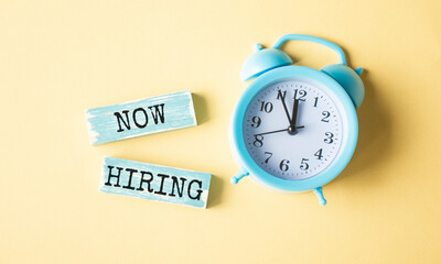 New Hiring text written on wood block. Now we are hiring text on the yellow table with a blue alarm clock, for your design.
