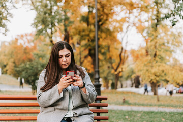 Beautiful young adult woman sitting on a bench in an autumn Park with a phone