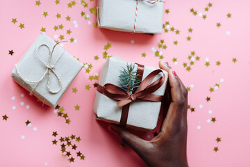 Hands of African woman holding present box with a bow on pastel pink background with stars and snowflakes confetti. Selective focus