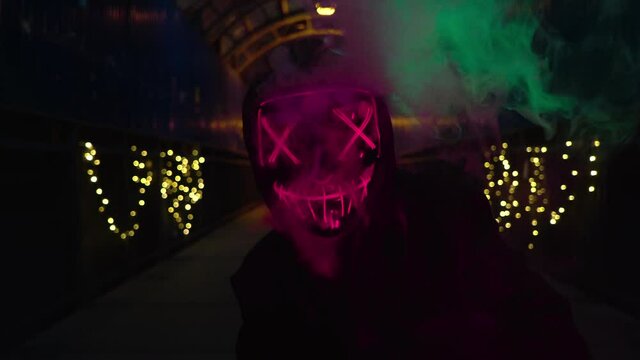 The man in the neon mask blows smoke. Thick green and pink smoke. Scary Halloween Purge killer costume.