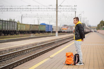 Fototapeta premium Young man stands on platform, waiting for train. Male passenger with backpacks on railroad platform in waiting for train ride. Concept of tourism, travel and recreation.