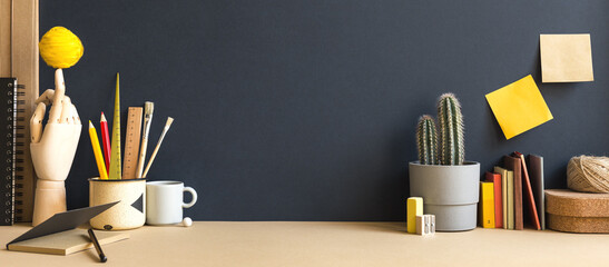 Creative desk with notebook, desk objects, office supplies, books, and cactus on a dark blue background.	
