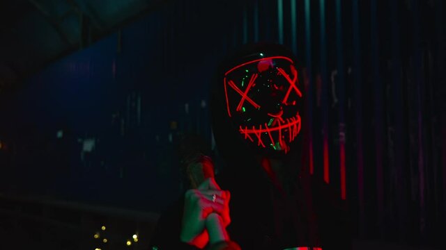The man in the neon mask blows smoke. Thick green and red smoke. Scary Halloween Purge killer costume.