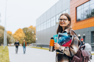 Fototapeta premium Portrait of girl student standing at university campus. Laughing Woman wearing braces, eyeglasses, bag with books having coffee break after lecture. Enjoying College life. Learning education concept.