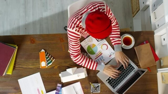 Top view of creative male designer in beanie hat and glasses working on project in office. He is looking at financial document and typing on laptop, then choosing color for house model exterior