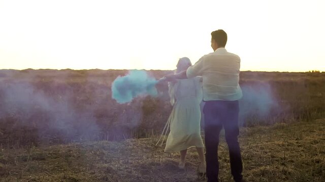 Gender Reveal Party With Caucasian Couple Who Spin Round Holding Hands With Smoke Bomb In Slowmo. Man And Pregnant Woman Are Expecting Baby Boy. They Are Dancing And Smiling In Summer Field On Sunset.