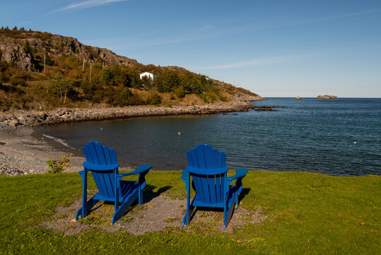 Two blue Adirondack chairs surrounded by grass overlooking the ocean and a small beach. The chairs are empty. The sky is blue with a little cloud near the horizon. The beach is sandy with no waves.