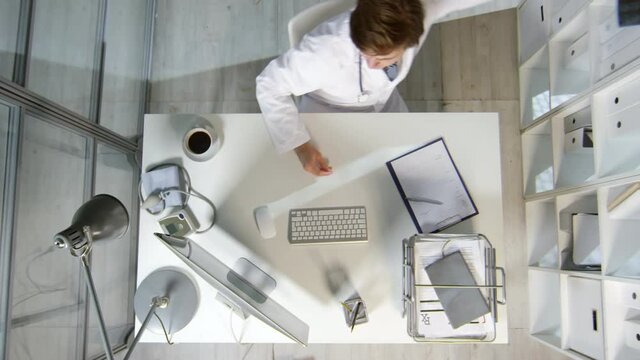 Directly Above Shot Of Male Doctor In White Coat Sitting At His Desk And Looking At MRI Of Patient, Then Leaving Office