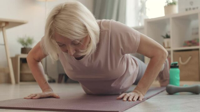 Going down from out of frame to POV of senior white-haired woman doing push ups on yoga mat in living room
