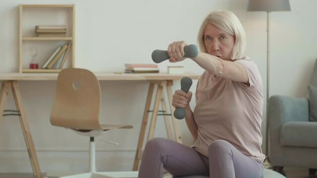 POV of elderly lively female gym coach hosting online class from home sitting and jumping on fitball doing punching moves holding small weights in hands looking and talking on camera