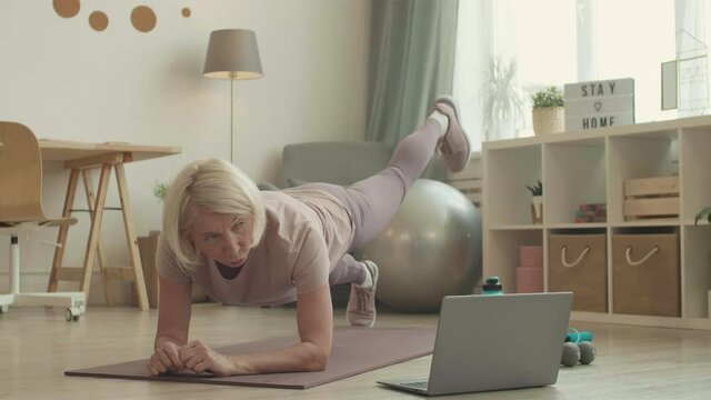 Experienced white-haired Caucasian fitness instructor providing online yoga class doing legs up plank exercise on floor at home in front of laptop
