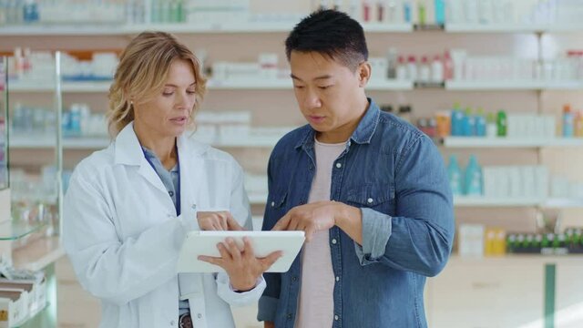 Close up woman and younger Asian intern cooperating in drugstore use tablet computer. Attracitve woman apothecarist talking with male colleague learning pills. Slow motion