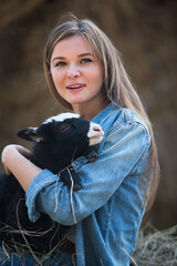 Naklejka na ściany i meble Young beautiful woman in a barn on the hay holding a small lamb