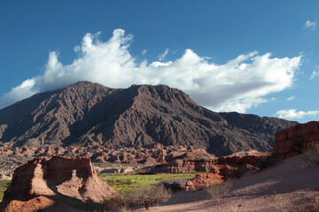 Magical view of the arid desert, green valley, red sand, sandstone formations and brown rocky mountains under a deep blue sky with beautiful clouds.  