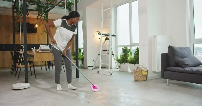 Beautiful african american woman mopping floor in living room with mop. Pretty housewife doing housework mopping while robotic hoover cleaning floor. Female concentrated on occupation. Daily routine.