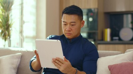 Serious asian young man use tablet computer sitting on sofa at home. Chinese businessman device handsome. Communication. Close up. Slow motion - Powered by Adobe