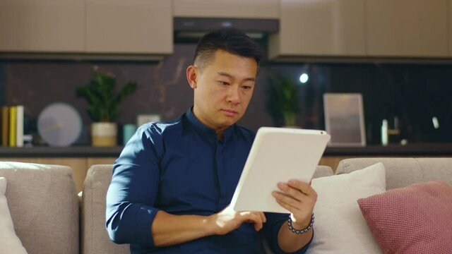 At home serious asian young man use tablet computer sitting on sofa. Chinese businessman device handsome. Communication. Close up. Slow motion