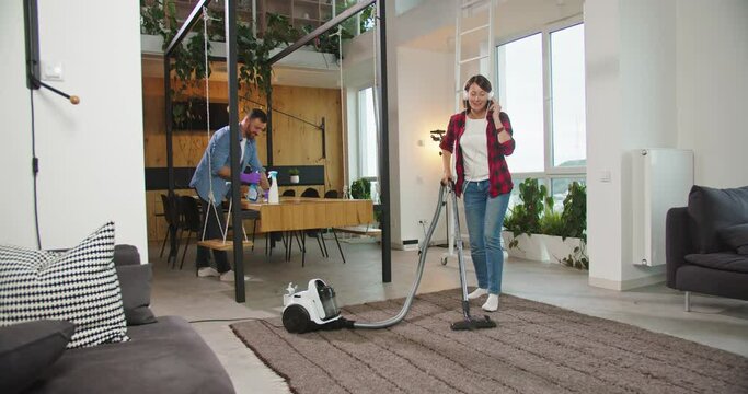 Happy couple clean-up home. Woman vacuuming floor, listening music in headphones and dancing, while man dusting table. Joyful couple cleaning apartment. Housekeeping, people and happiness concept.