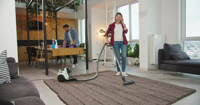 Happy couple clean-up home. Woman vacuuming floor and speaking using smartphone, while man dusting table. Joyful couple cleaning apartment, household and housekeeping.