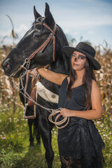 Obraz premium Girl disguised as Catrina (Mexican female character of death) dressed in black, in the middle of a corn field, with mirasoles flowers, stroking a horse 13
