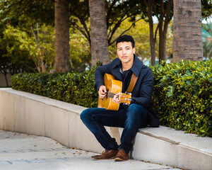 Obraz premium Young student sitting on the street in the city public park while playing guitar - street musician, student off-campus life, autumn break time