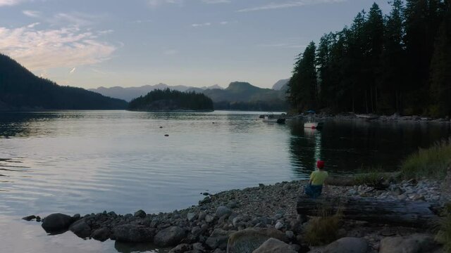 Woman Sitting On A Tree Log Enjoying The View Of Sechelt Inlet During Sunset On The Sunshine Coast Near Egmont, British Columbia, Canada. - aerial pullback shot