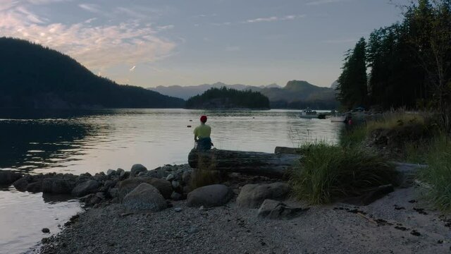 Woman Sitting On A Tree Log Enjoying The Beautiful Sunset Views of Sechelt Inlet, A Pacific Ocean Fjord In Sunshine Coast Near Egmont, British Columbia, Canada - drone pullback shot (ascending)