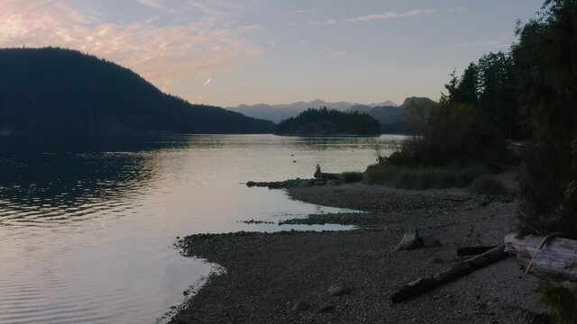 Glorious Sunset By The Sechelt Inlet In Sunshine Coast Near Egmont, BC, Canada With A Lady Sitting On The Shore - descending drone shot