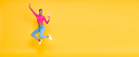 Fototapeta na wymiar Smiling energetic African American woman wearing headphones listining to music from smartphone and jumping on isolated yellow banner background with copy space