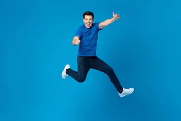Tuinposter Smiling handsome American man joyfully jumping and doing double thumbs up gesture isolated on blue studio background © Atstock Productions