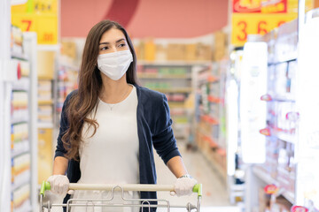 Beautiful woman wearing medical face mask and rubber glove push shopping cart in supermarket department store. girl looking at grocery shelf. new normal lifestyle during Coronavirus or covid pandemic