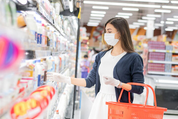 Beautiful woman wearing medical face mask and rubber glove with grocery trolley picking up daily milk on product shelf. shopping at supermarket in new normal lifestyle concept during Coronavirus.