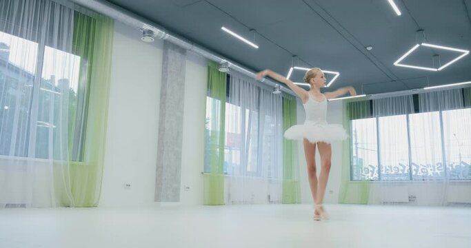 Cute little ballerina in white suit with tutu performs classical dance in light studio with large windows