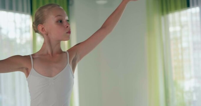 Concentrated little girl in white suit dances doing classical ballet spins at window in studio closeup slow motion