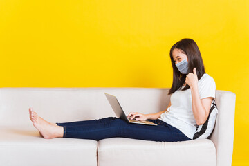 Fototapeta premium Portrait Asian of beautiful young woman sitting on sofa wearing medical face mask protective she work from home with laptop computer during Coronavirus studio shot isolated on yellow background