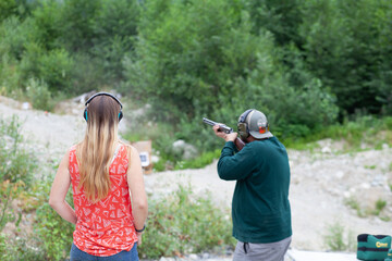 A young women stands behind a man holding a shotgun downrange, shooting clay pigeons to practice...